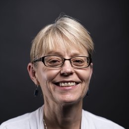 lady with blonde hair pulled back in ponytail wearing glasses with, grey shirt and white cardigan in front of black background