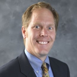 Man with dark blonde hair wearing black suite and blue button up shirt with gold paisley tie