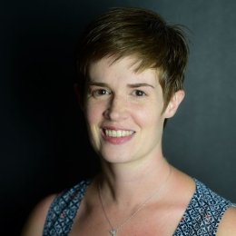 Lady with short pixie cut brown hair wearing sleeveless top and necklace in front of grey background