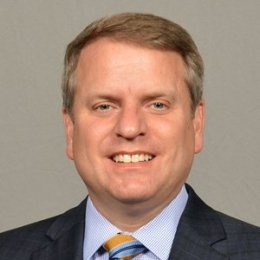 Greg Mason headshot, man wearing dark suit with blue button up shirt, and striped tie in front of grey background
