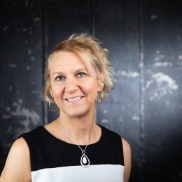 Lady with blonde hair in ponytail wearing black and white top standing in front of black backdrop