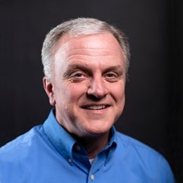 Man with short grey hair with blue button up dress shirt in front of black background