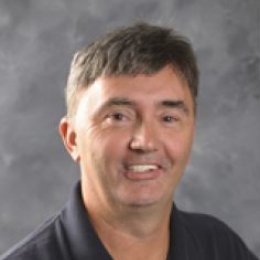 Man with short dark hair wearing a black polo shirt in front of grey background