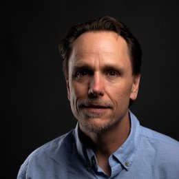 Man with dark hair wearing blue button up in front of dark background