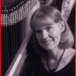 Lady with blonde shoulder length hair wearing dark top and necklace sitting next to a harp