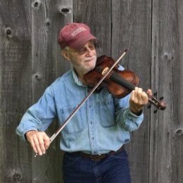 Man wearing red ball cap and denim button up shirt playing fiddle in front of barn