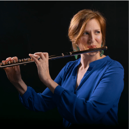 Lady with auburn hair with royal blue top playing the flute in front of black background
