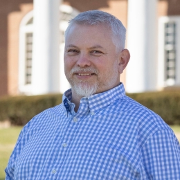 A man poses for a photo in front of a brick building with white columns.