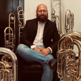 Man with beard and glasses wearing black sport coat with white button up and jeans sitting next to trombones and tubas