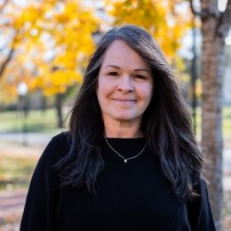 Lady with long dark hair wearing black sweater and necklace standing in front of yellow fall tree on campus