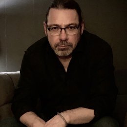 Man with dark hair and glasses wearing black button up shirt in front of dark wall