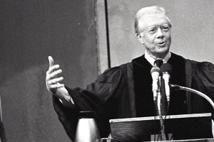 Former President Jimmy Carter spreads his arms wide as he speaks behind a podium.