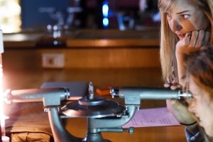 One female student watches as another looks through a scientific tool.