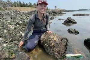 A young woman kneels behind a large stone where a rocky beach meets the ocean.