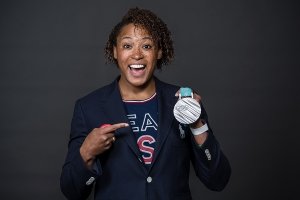 A smiling woman holds a silver olympic medal in one hand while pointing to it with her other hand.