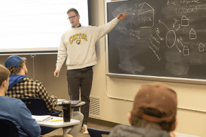 Derek Nafziger wears a Centre College sweatshirt as she stands at a black chalkboard teaching students about real estate insurance.