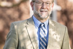 A man wearing a tan sports coat and blue shirt and tie poses for a photo in front of a brick building.