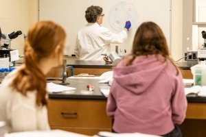 One man and two women watch as a professor wearing a white lab coat writes on a white board.