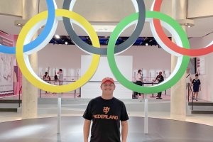 A man wearing a t-shirt and baseball cap poses in front of the Olympic Rings.