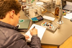 We see a young man writing on a piece of paper placed on a lab table alongside scientific instruments.