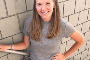A woman wearing a grey t-shirt smiles for the camera while posing against a tile wall.