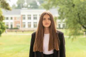 A young woman with long brown hair poses for a photo with a green field and brick buildings in the background.