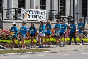 Nine college students wearing matching blue t-shirts pass a banner hanging from a downtown building that states &quot;Welcome Centre Students.&quot;
