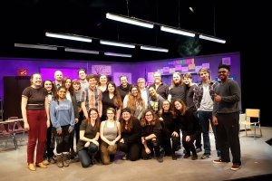 A group of student actors, along with faculty members post for a photo on stage in front of a set designed to look like a high school classroom.