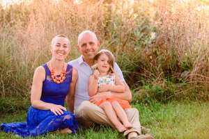 An image of Megan Garrison sitting with her husband and daughter in the grass with tall grass and plants behind them.