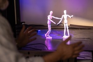Two posable miniature figures are seen on a model stage as a man's hands gesture in the foreground.