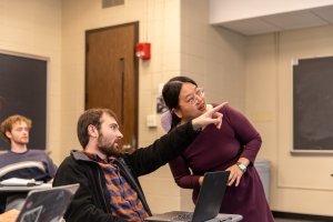 A student points to something projected out of the frame as a professor looks in the direction he is indicating.