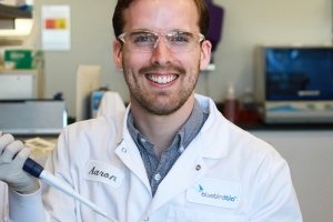 A man wearing a white lab coat holding scientific instruments smiles for a photo.