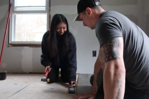 A student and staff member are working together on a flooring project in North Carolina. The student is kneeling, holding a hammer and flooring tool, while the staff member leans forward to assist.