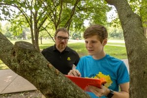 A young man in a blue t-shirt writes on a clipboard while standing near a tree as another man looks on.