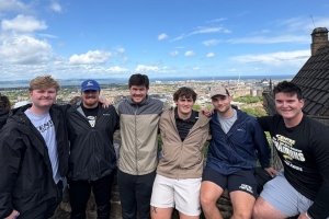 A group of 6 men smile for a photo. They are on a roof of a building with a view of a village behind them.