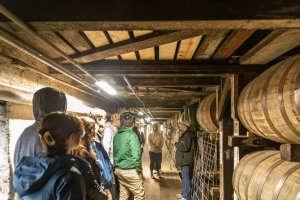Students listen as to a man as they all stand surrounded by large wooden barrels in a bourbon aging warehouse, known as a rickhouse.