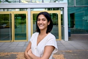 Mehak Mittal is smiling and standing in front of doors to a building, posing for a headshot.