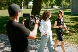 A man with a large camera videos two women walking and talking along an outdoor pathway.