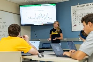 A young woman speaks with follow students seated in a classroom as a line graph is shown on a screen behind her.
