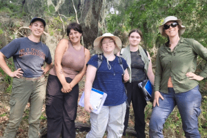 A group of students and faculty are smiling while wearing gear for an Archaeological dig.