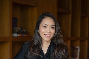 A woman ina. dark blazer and black button-up shirt poses for a photo in front of a wall of wooden shelving
