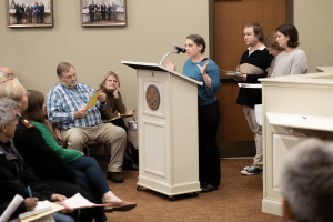 A woman speaks into a microphone at a lectern in Danville City Hall as students stand behind her and a seated audience looks on.