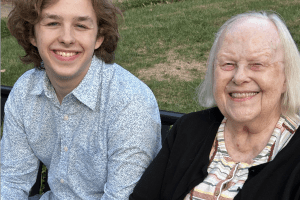 A college-aged man in a blue shirt sits outside next to his grandmother as both smile for the photographer.