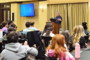 A professor is sitting at the front of the room, presenting to a group of students.