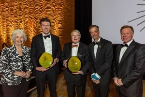 One woman stands alongside four men in tuxedos, two holding gold and black Associate of the Year awards.