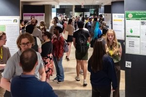 A hallway is filled with people talking with one another in front of research posters mounted along the length of the hallway.