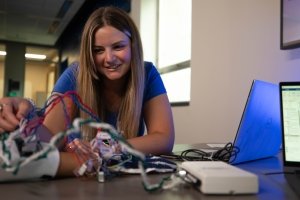 Madison Bates adjusts a device with man wires attached to it that is strapped to the hand of a person off camera as a computer screen shows data.