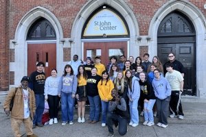A group of more than 20 young men and women, many wearing Centre College clothing, pose for a photo on the step of a brick building, upon which a sign reads "St. John Center."