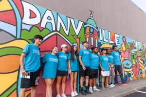 Nine students wearing identical t-shirts pose in front of a colorful mural that says "Danville."