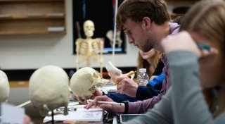 Students holding skeleton skulls in a forensics lab with a full skeleton in the background